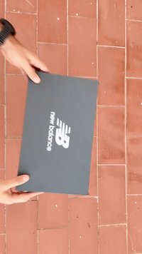 Szürke karton cipődobozt tartanak kézben, amelynek fedelén fehér "new balance" logó látható, mögötte csempézett padló.