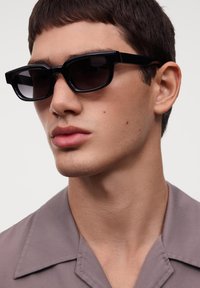 Melnas taisnstūra formas saulesbrilles ar biezām rāmēm un tumšiem stikliem, ko valkā vīriešu modelis gaiši pelēkā krekla fona priekšā.