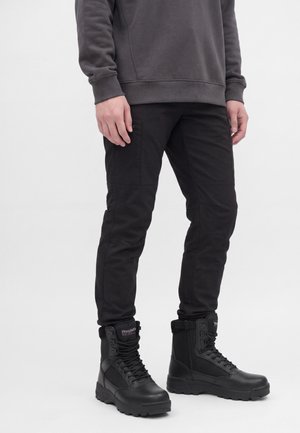 Persona vistiendo pantalones cargo negros y botas negras altas con cordones y cremalleras laterales, de pie frente a un fondo blanco.