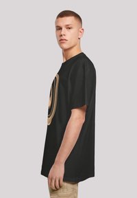 Czarny bawełniany t-shirt z dużym, okrągłym, abstrakcyjnym wzorem w odcieniu złota. Luźny krój, krótkie rękawy, klasyczny okrągły dekolt.