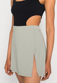 Saia cinza-claro com uma fenda alta, feita de material suave. Apresenta uma cintura ajustada e costuras limpas. Combinada com um bodysuit preto sem mangas.
