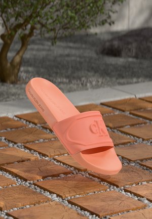 Einzelne korallenrosa Calvin Klein Slide-Sandale schwebt über quadratischen Holzdielen mit kleinen Kieselsteinen im Freien in der Nähe eines Baumes.