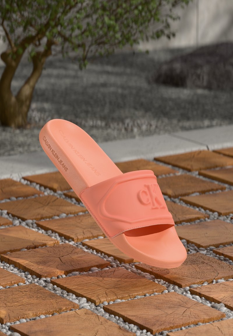 Sandale à glissière corail rose unique Calvin Klein flottant au-dessus de carreaux carrés en bois avec de petits cailloux à l'extérieur près d'un arbre.
