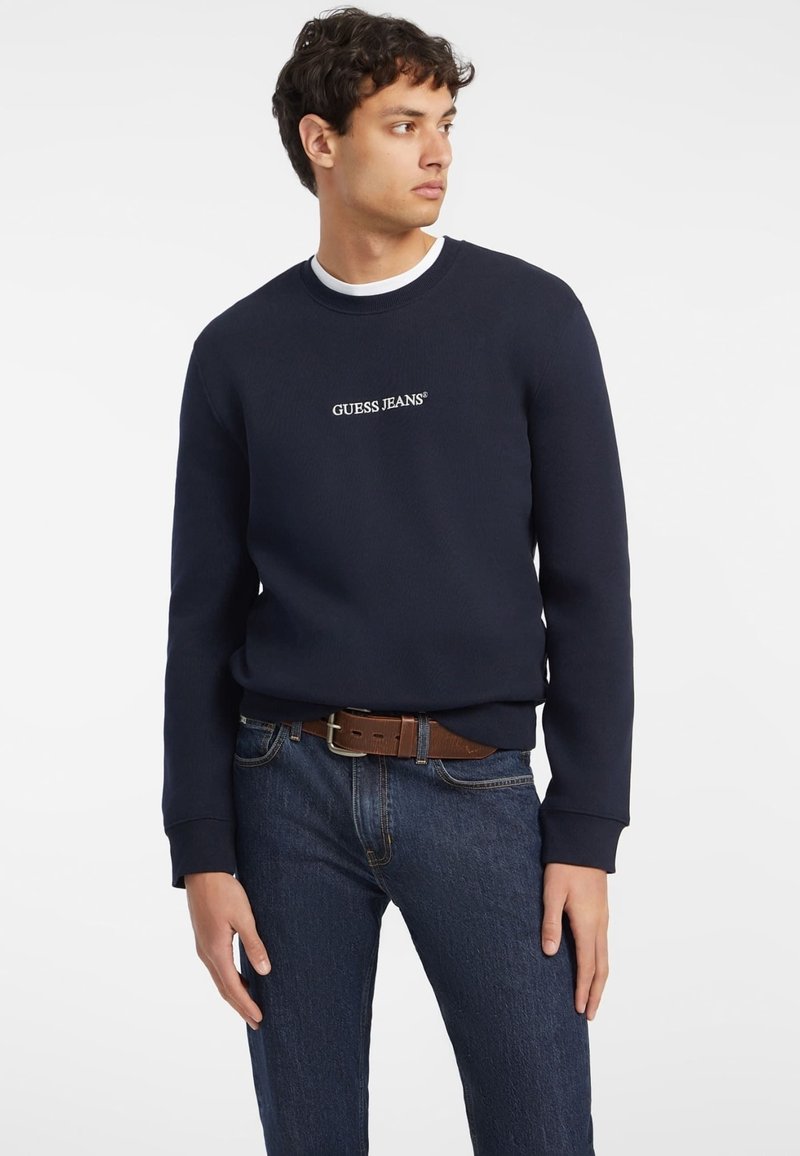 Sötétkék pulóver bordázott nyakkal és mandzsettával, középen fehér "GUESS JEANS" felirattal; sötét farmernadrággal és barna övvel párosítva.