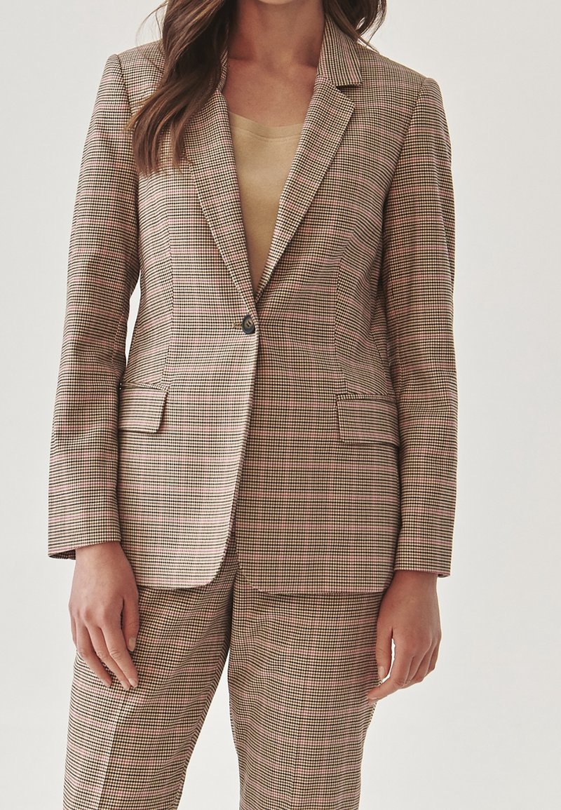 Femme portant un blazer à petits carreaux beige et marron et un pantalon assorti avec un haut beige clair en dessous.