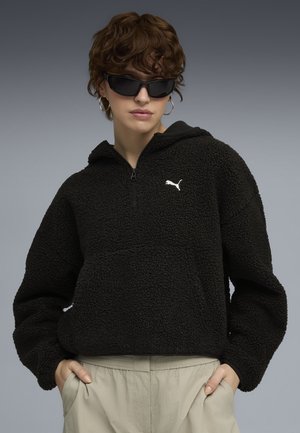 Persona con capelli corti e ricci che indossa una felpa nera con zip e logo Puma, occhiali da sole scuri, orecchini a cerchio e pantaloni beige con le mani nelle tasche.