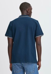 Moški obrnjeni stran nosi temno modro kratkouhanovno polo majico z diagonalnim ovratnikom in modre kavbojke proti enobarvni ozadju.