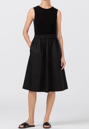 Femme portant un haut noir sans manches, une jupe noire mi-longue avec une ceinture élastique et des sandales noires à enfiler sur un fond clair uni.