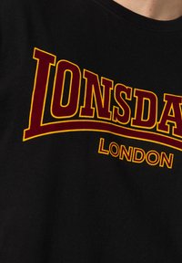 Tricou negru din bumbac, cu un text îndrăzneț în culorile roșu și galben care spune "LONSDALE LONDON" într-un format curbat.