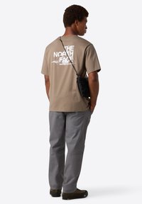 Mies, joka kääntää selkänsä, päällä beigestä The North Face -t-paidasta, harmaista housuista, ruskeista kengistä ja pienestä mustasta olkalaukusta.