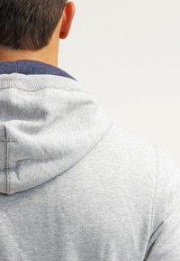 Grijze hoodie van zacht materiaal, met een marineblauwe kraag. Zichtbare stiksels en een trekkoord in de capuchon creëren een ontspannen ontwerp.