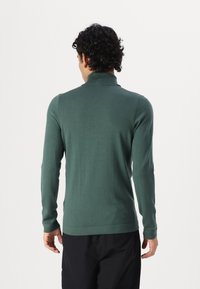 Pull vert en maille avec un col montant, des manches longues et une coupe ajustée au tissu lisse, porté avec un pantalon noir.