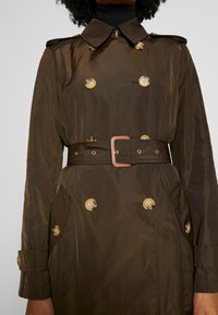 Mörkbrun trenchcoat med bälte och dubbelknäppning med sköldpaddsmönstrade knappar, bärs över en svart polotröja.