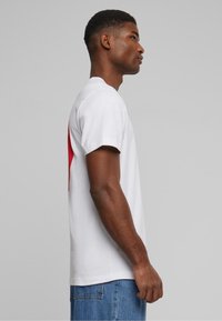 Hvit bomulls T-skjorte med et rødt abstrakt design på baksiden. Korte ermer og rett snitt. Brukt med blå denimjeans.