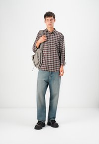 Checkered brown and white button-up shirt, loose light blue jeans, black shoes, and a gray shoulder bag with zippers. Simple styling.