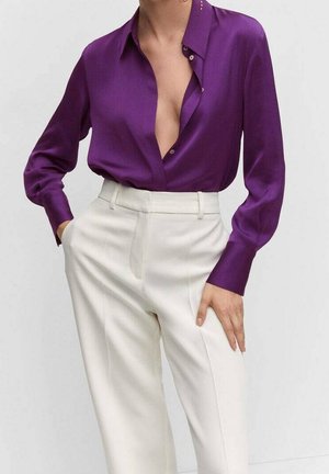 Frau trägt eine dunkelviolette Seidenbluse mit tiefem Ausschnitt, die in eine hoch taillierte weiße, maßgeschneiderte Hose gesteckt ist, eine Hand in der Tasche.