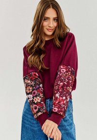 Burgundowa bluza z rękawami w kwiatowy wzór; posiada ściągacze przy mankietach, okrągły dekolt i miękki materiał; zestawiona z jasnoniebieskimi dżinsami.