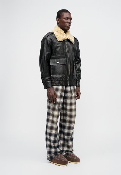 Veste en cuir noire avec un col en shearling beige, deux poches avant, associée à un pantalon à carreaux à large jambe et des chaussons à motifs marron.