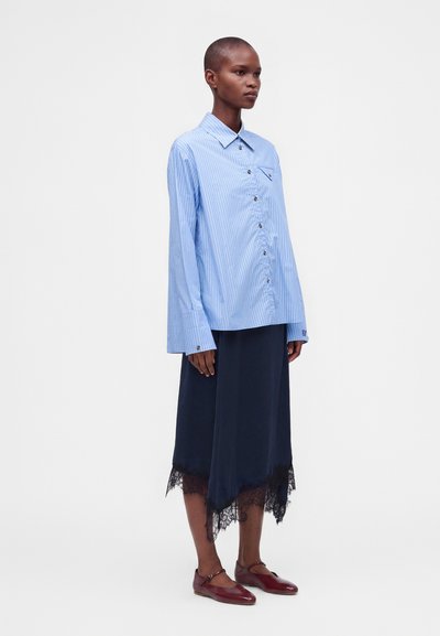 Femme portant une chemise rayée bleue à boutons, une jupe bleu marine bordée de dentelle et des chaussures Mary Jane bordeaux, debout sur fond blanc.