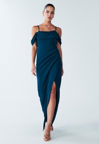 Teal avondjurk met off-shoulder mouwen, aansluitende lijfje, gedrapeerde rok en hoge split. Zachte stof met een gladde textuur. Sandalen met gouden accenten.