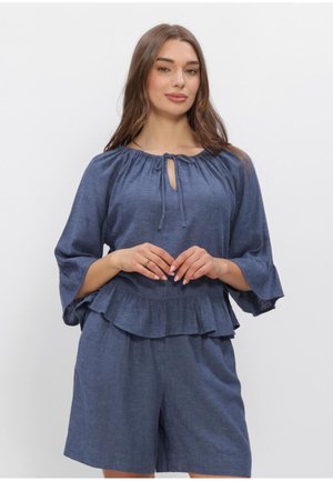 Femme aux longs cheveux bruns portant un chemisier bleu à volants avec un col à nouer et un short bleu assorti, debout devant un fond clair uni.