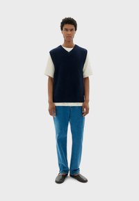 Gilet tricoté bleu marine avec un col en V sur un t-shirt blanc, associé à un jean bleu clair et des chaussures noires à enfiler.