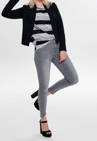 Femme portant une chemise rayée grise et noire, une veste noire, un jean skinny gris et des sandales à talons hauts noires, posant avec une jambe pliée.