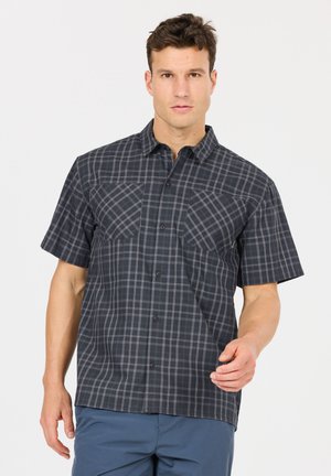 Mann trägt ein dunkelgraues, kurzärmeliges kariertes Hemd mit zwei Brusttaschen und blaue Hosen, steht vor einem weißen Hintergrund.