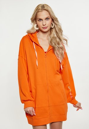 Vestito con zip arancione, dotato di una grande tasca frontale, cappuccio con cordino e maniche oversize. Ricamo testuale sulla manica. Tessuto morbido.