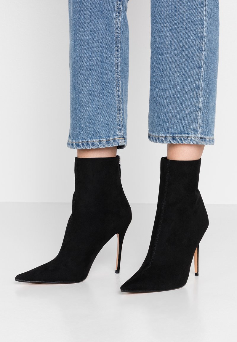 Bottes cheville en daim noir, à bouts pointus et talons stilettos hauts. Présentent une surface lisse et une fermeture zippée à l'arrière.