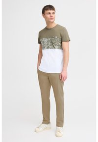 Grün-weißer Farbblock-T-Shirt mit strukturiertem Design, kombiniert mit beigen Hosen und weißen Sneakern. Das T-Shirt verfügt über eine Brusttasche.