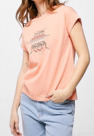 Mujer con camiseta color melocotón con un diseño de escarabajo estampado y vaqueros azul claro, una mano en el bolsillo y la otra relajada a su lado.