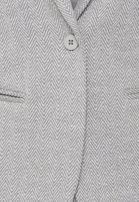 Blazer gris à motif chevrons avec un seul bouton rond et deux poches horizontales à l'avant.
