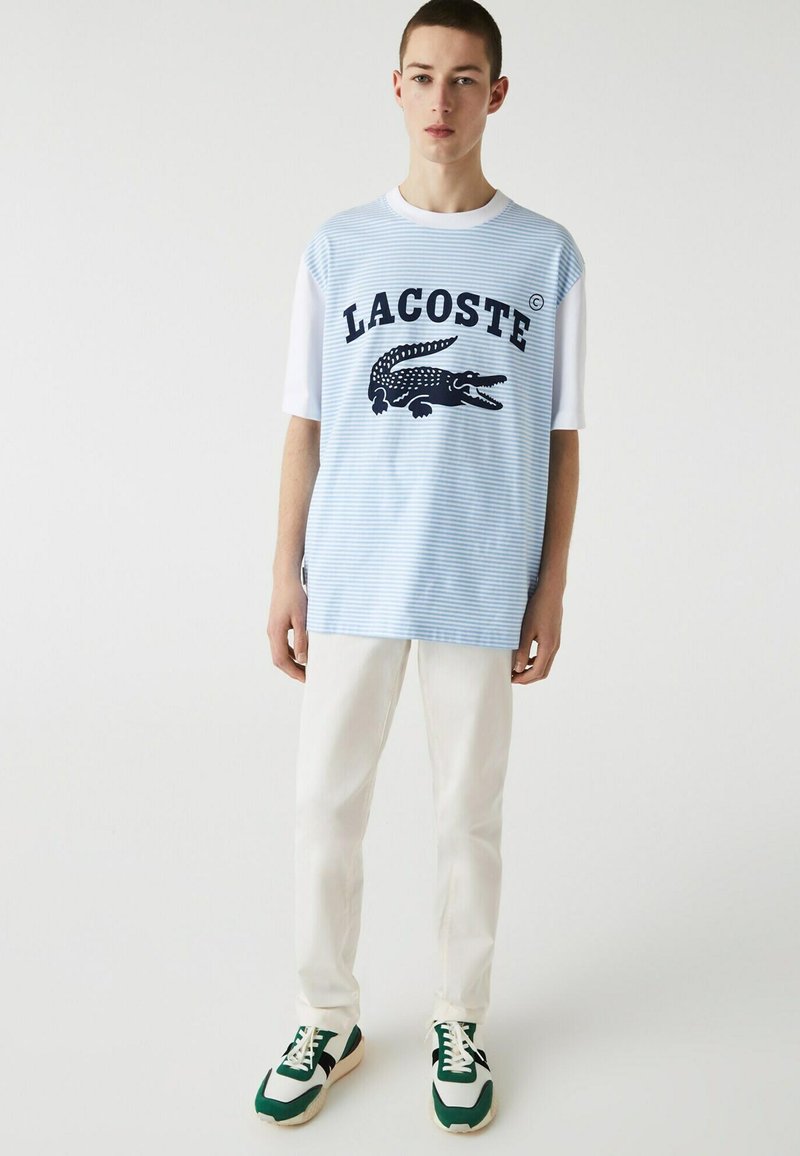Camicia a maniche corte Lacoste con righe azzurre, caratterizzata da un logo di coccodrillo nero e testo. Indossata con pantaloni bianchi e sneakers con dettagli verdi.