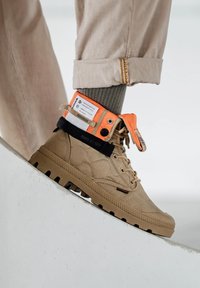 Sneaker alte beige in materiale di tela testurizzato, suola in gomma, con dettagli arancioni e neri e un'etichetta visibile sulla caviglia.