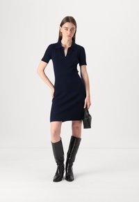 Abito a maglia a coste blu navy con maniche corte, colletto e fit slim, abbinato a stivali neri lucidi al ginocchio e una piccola borsa nera.