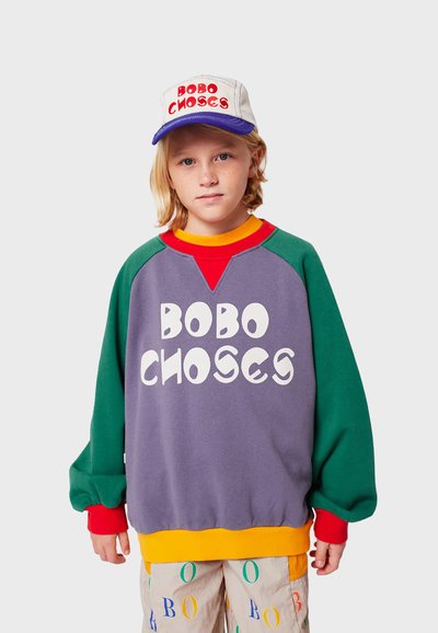Felpa multicolore con corpo viola, maniche verdi e dettagli rossi; grande testo bianco "BOBO CHOSE." Abbinata a un cappellino.