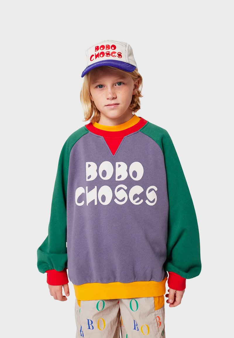 Felpa viola oversize con maniche verdi, orlo giallo e testo bianco in grassetto "BOBO CHOSES." Abbinata a un cappellino grigio con lettering rosso.