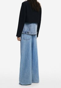 Gekürzter schwarzer Blazer über einer hellblauen, geschichteten Denimjacke, kombiniert mit weit geschnittenen hellblauen Denimhosen, die Gesäßtaschen und eine weiche Textur aufweisen.