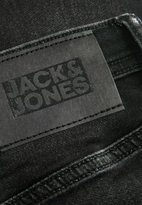 Melna denima auduma, kuram ir etiķete ar "JACK & JONES" uzrakstu glancētā apdarē. Šuvju krāsa ir melna, un kopējā virsma ir gluda.