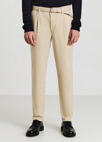 Pantaloni beige su misura con una texture liscia, pieghe frontali e una cintura. Abbinati a scarpe nere e a un top lungo scuro.