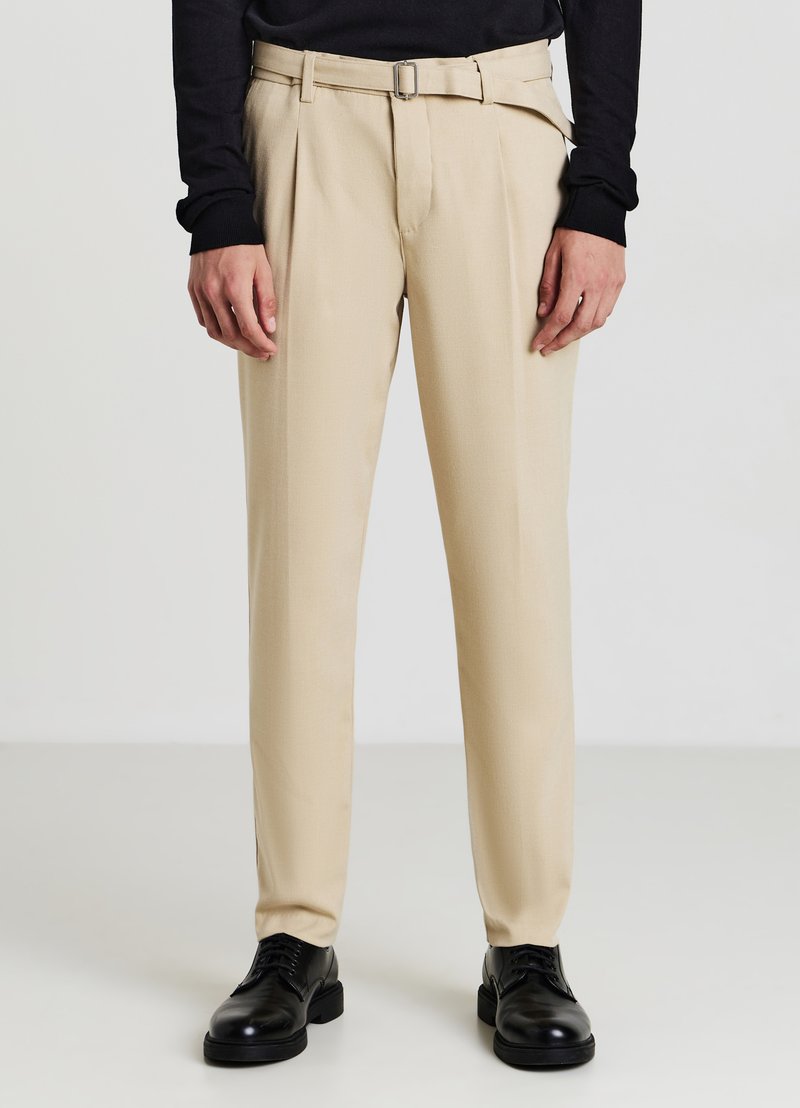 Pantaloni beige su misura con una texture liscia, pieghe frontali e una cintura. Abbinati a scarpe nere e a un top lungo scuro.