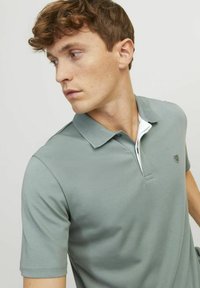 Šedé polokošile s krátkými rukávy, texturovaným límcem a bílými akcenty na krku. Na hrudi má malé logo. Hladká látka.