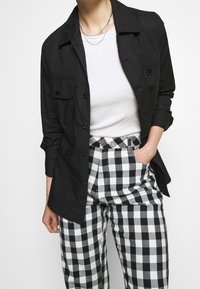 Camicia leggera nera con bottoni e due tasche sul petto, indossata sopra una maglia bianca aderente e abbinata a pantaloni a vita alta a quadretti bianco e neri.
