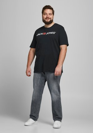 Schwarzes T-Shirt mit kurzen Ärmeln und "JACK & JONES"-Logo, kombiniert mit hellgrauen Jeans und weißen Sneakers, vor einem einfarbigen Hintergrund stehend.
