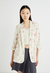 Blazer imprimé floral en crème clair, avec des manches retroussées, une coupe décontractée et des fleurs pastel roses et bleues sur l'ensemble.