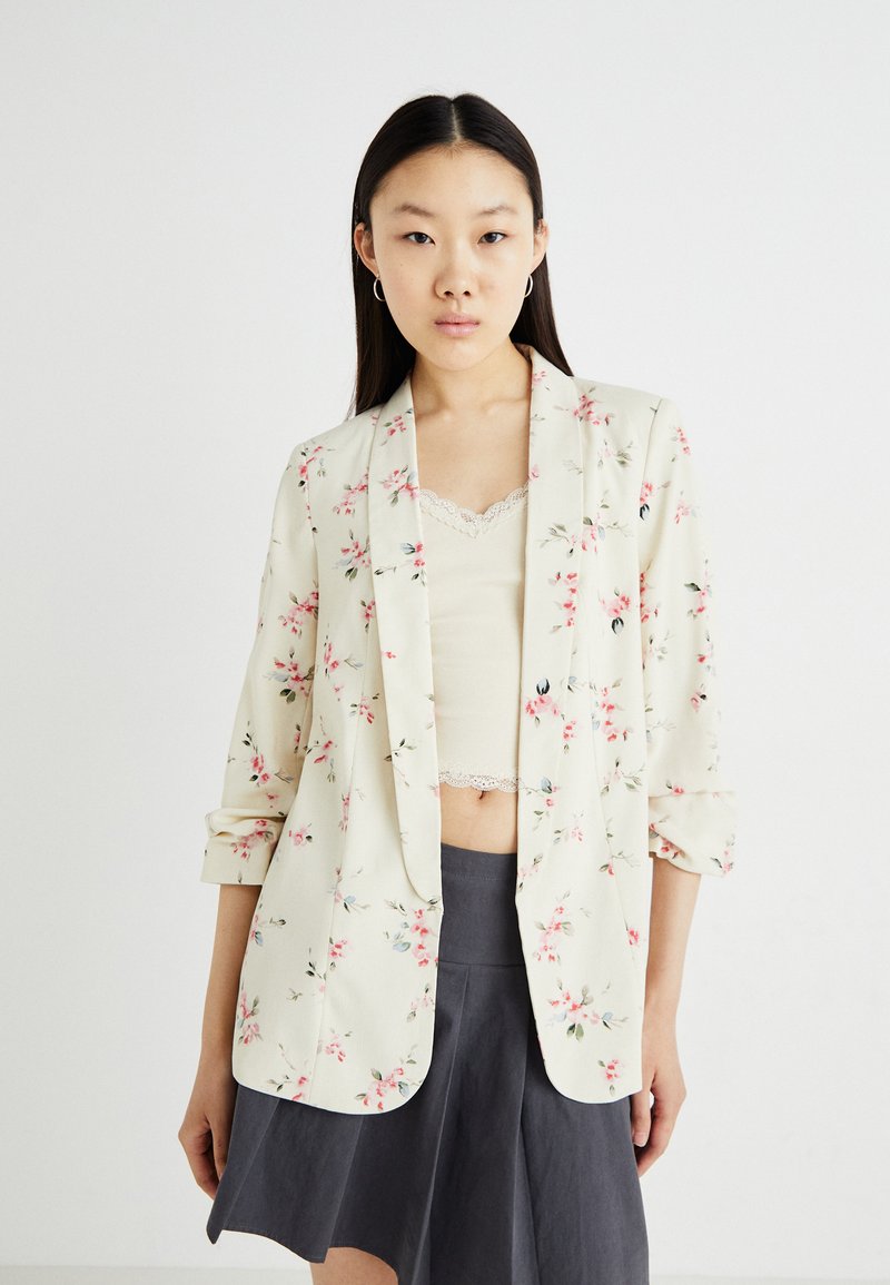 Blazer imprimé floral en crème clair, avec des manches retroussées, une coupe décontractée et des fleurs pastel roses et bleues sur l'ensemble.