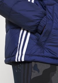 Marinblå puffjacka med vit tre-strips design på sidan. Har elastiska muddarna och en zip-försedd ficka på framsidan.