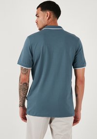 Moška polo majica temno teal barve s kratkimi rokavi, belim ovratnikom in poudarki na rokavih, gladka tkanina ter majhnim logotipom na strani.