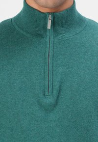 Mann trägt einen türkisfarbenen, gerafften Halbreißverschluss-Pullover mit silbernem Reißverschluss-Zug, markiert mit "Tom Tailor", zu sehen sind Ausschnitt und oberer Brustbereich.